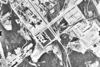 A imagem é uma fotografia aérea em preto e branco de uma área urbana e rural, com grandes extensões de terreno. Diversas estradas e caminhos sinuosos são visíveis, cortando a paisagem. Há áreas com densa vegetação escura, contrastando com clareiras e terrenos abertos. Várias estruturas construídas, incluindo edifícios e complexos de edifícios, podem ser observadas em diferentes partes da imagem. Há também o que parecem ser grandes campos abertos ou pistas, indicando possíveis infraestruturas.