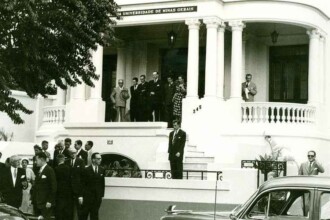 A imagem, em preto e branco, mostra um grupo de homens em frente a um edifício de dois andares com uma varanda. O prédio tem uma fachada branca com colunas. No topo da fachada, o texto "UNIVERSIDADE DE MINAS GERAIS" é visível. Há duas escadarias que levam à entrada principal, e um homem está parado na parte mais alta da escada. Um grupo de homens vestidos com ternos escuros está reunido na parte de baixo. À direita, um carro antigo está parcialmente visível. A vegetação de uma árvore está no canto superior esquerdo.