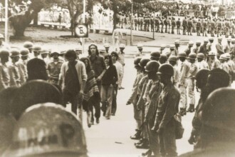 A imagem em preto e branco captura uma cena com múltiplos grupos de pessoas em um espaço aberto, que parece ser um pátio ou uma grande praça. Em primeiro plano, a parte superior de capacetes, possivelmente militares ou de segurança, está visível, indicando a perspectiva de quem observa a cena. No centro da imagem, há uma fileira de indivíduos em pé, de frente para a câmera, alguns com boinas ou capacetes. Mais ao fundo, à direita, uma longa fila de pessoas, algumas vestindo o que parecem ser uniformes militares e capacetes, está organizada em formação. À esquerda do centro, um grupo menor de pessoas, vestindo roupas civis e com expressões sérias ou atentas, caminha em direção ao observador. Ao fundo, árvores e um sinal de trânsito de "Estacionamento Proibido" são visíveis, com mais pessoas em segundo plano. O céu é claro e a iluminação da cena é uniforme.