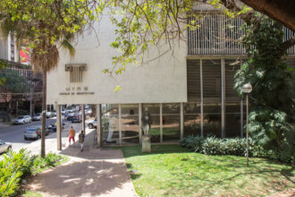 A imagem mostra a fachada da Escola de Arquitetura da UFMG, um edifício de cor clara com grandes janelas de vidro que refletem o ambiente externo. Na parte superior da fachada, um emblema escuro e as letras "UFMG ESCOLA DE ARQUITETURA" são visíveis. Em frente ao prédio, há uma estátua escura em um pedestal de pedra, ladeada por arbustos verdes. Um caminho pavimentado leva à entrada do edifício, e no lado esquerdo da imagem, uma rua com carros estacionados e algumas pessoas caminhando pode ser vista. Árvores com folhagem verde emolduram a cena, com uma proeminente no canto superior direito, e o céu azul com algumas nuvens é visível na parte superior.