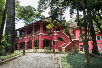 A imagem mostra uma grande casa de dois andares pintada em um tom vibrante de rosa avermelhado, cercada por uma densa vegetação. A casa possui um telhado de telhas escuras, varandas com pilares brancos e janelas envidraçadas no andar superior. No lado direito da fachada, uma escadaria externa com corrimãos brancos e detalhes em arco leva ao andar superior. Em primeiro plano, um caminho de paralelepípedos serpenteia em direção à casa, ladeado por áreas ajardinadas com grama baixa e plantas. Várias árvores altas com troncos largos e folhagem verde escura emolduram a casa, algumas delas com cipós pendurados. Há também postes de madeira baixos ao longo do caminho. O céu é visível em pequenas aberturas entre as folhas das árvores, indicando um dia claro.