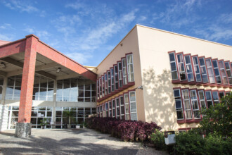 A imagem mostra um edifício de arquitetura moderna, com fachada de cor bege claro e detalhes em tons de bordô ou terracota. No lado esquerdo da imagem, uma estrutura de entrada com colunas largas na cor terracota e um telhado plano se estende sobre uma grande área envidraçada, que serve como a entrada principal do prédio. À direita da entrada, o corpo principal do edifício apresenta várias janelas retangulares com molduras escuras e persianas internas, dispostas em fileiras horizontais e em um padrão inclinado que acompanha a inclinação do telhado. Um canteiro com arbustos de folhagem escura e algumas plantas menores se estende ao longo da fachada. O céu azul com nuvens brancas, e algumas sombras de árvores, completam a cena.