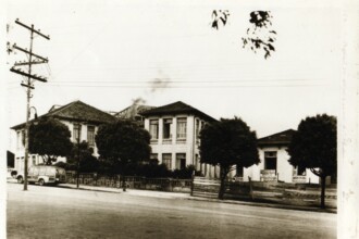 A imagem em preto e branco retrata uma paisagem urbana com vários edifícios e vegetação. À esquerda, um poste de madeira com múltiplos fios e isoladores se eleva verticalmente. Abaixo dele, na rua, um veículo de passageiros, semelhante a uma van antiga, está estacionado. O centro da imagem é dominado por uma série de edifícios com telhados inclinados de telhas escuras. O prédio mais à esquerda e o central são de dois andares, com janelas retangulares. No prédio central, é possível ver uma coluna de fumaça subindo ao fundo. Árvores com folhagem densa estão espalhadas em frente e entre os edifícios, obscurecendo parte das fachadas. O primeiro plano mostra uma rua pavimentada.