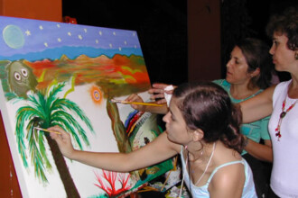 A imagem colorida mostra três mulheres engajadas na pintura de uma tela grande, disposta em um cavalete. A tela exibe uma paisagem vibrante e estilizada, com elementos como um sol radiante, montanhas em tons de laranja e marrom, um céu azul com uma lua e estrelas, e uma palmeira proeminente no primeiro plano esquerdo. A mulher mais próxima do espectador está focada na pintura da palmeira, segurando um pincel. Atrás dela, uma segunda mulher também está ativamente pintando na tela. Uma terceira mulher, com cabelos cacheados e usando um colar de contas, observa a pintura com atenção, posicionada mais à direita. O ambiente parece ser interno, com iluminação artificial, e a atmosfera sugere um evento ou atividade criativa.