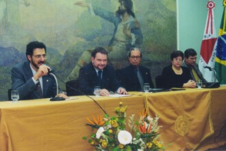 A imagem mostra cinco autoridades sentadas atrás de uma longa mesa conduzindo o evento. A pessoa à esquerda, com óculos e barba, segura um microfone. Todas as cinco pessoas estão vestidas com trajes formais. Ao fundo, uma grande pintura de parede exibe uma cena histórica com uma figura central em traje militar e bandeiras, incluindo a bandeira do Brasil e a bandeira do estado de Minas Gerais, estão visíveis no lado direito da imagem.