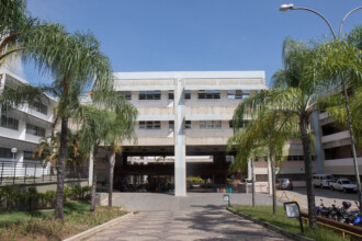 A imagem mostra um grande edifício com uma arquitetura que remete a meados do século XX, com múltiplos andares e uma predominância de concreto e janelas horizontais. Há uma estrutura central que cria uma passagem ou pátio coberto, por onde se pode ver o interior do campus. A área em frente e ao redor do prédio é ajardinada, com palmeiras altas em primeiro plano e outros arbustos. Há um caminho pavimentado que leva à entrada central. Veículos, incluindo carros e motos, estão estacionados na lateral direita da imagem. O céu é azul e claro, indicando um dia ensolarado.