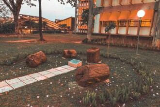 A imagem mostra um jardim, com um caminho de lajotas claras que se estende pelo gramado verde. O caminho é ladeado por troncos de árvores cortados, dispostos como assentos informais. O gramado é decorado com pequenas plantas e flores dispersas. No fundo da imagem, um edifício grande e moderno, com fachada amarela e janelas retangulares, é visível. À direita do caminho, um poste alto com uma luminária esférica ilumina a cena, sugerindo que a foto foi tirada ao entardecer ou em um dia com pouca luz. O céu está ligeiramente visível no topo, com tons de azul e roxo.