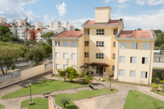 A imagem mostra um edifício de quatro andares, de cor bege claro, com telhados de telha colonial vermelha. As janelas do edifício possuem persianas brancas, algumas abertas e outras fechadas. Uma pequena marquise de telha protege a entrada principal. No topo do edifício, há uma estrutura retangular vertical e antenas. Em frente ao edifício, um gramado com caminhos de pedra serpentina e áreas verdes. Ao longo dos caminhos, postes de luz baixos estão visíveis. No lado esquerdo, uma parte de um muro baixo e algumas árvores, com uma rua e outros edifícios ao fundo, compõem o cenário. O céu é azul com nuvens brancas, e a iluminação geral sugere um dia claro.