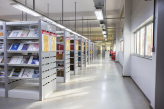 A imagem mostra o interior de uma biblioteca com longas fileiras de estantes de livros brancas e cinzas, dispostas paralelamente ao corredor central. As prateleiras estão parcialmente preenchidas com livros e pastas, alguns exibindo suas capas. Sinalizações verticais em cores diferentes indicam as seções, como "PERIÓDICOS". O ambiente é iluminado por longas luminárias fluorescentes no teto branco, que se estendem por todo o comprimento do corredor. No lado direito, há uma parede branca com várias janelas retangulares que permitem a entrada de luz natural. Ao fundo, uma pessoa está sentada em uma cadeira vermelha, utilizando um computador em uma mesa. O chão é claro e liso, refletindo a luz do teto.