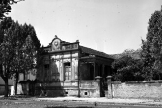 A imagem em preto e branco da década de 1960 apresenta uma antiga casa de arquitetura ornamentada, que serviu como escola de arquitetura da Universidade de Minas Gerais (UMG). A casa, com detalhes esculpidos na fachada, janelas de madeira fechadas e uma varanda coberta com grades de ferro, domina o centro da imagem. Um muro de pedra baixo e um portão de ferro delimitam a propriedade à direita, enquanto uma grande árvore com folhagem densa se destaca à esquerda.