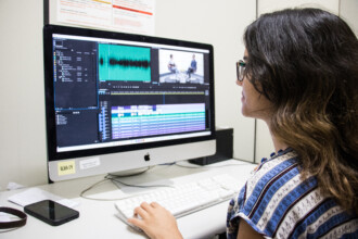 A imagem mostra uma pessoa sentada em frente a um computador iMac, de perfil, focada na tela. A pessoa usa óculos e tem cabelos longos e escuros. Ela veste uma blusa estampada com padrões em tons de azul e branco, e suas mãos estão posicionadas sobre um teclado branco, enquanto uma das mãos segura um mouse também branco. A tela do computador exibe um software de edição de vídeo, com múltiplas trilhas de áudio e vídeo, gráficos de ondas sonoras e uma pré-visualização de duas pessoas sentadas conversando. Ao lado esquerdo do monitor, há um telefone celular e o que parece ser uma pulseira ou acessório, sobre a mesa branca.