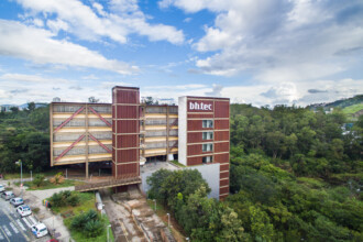 A imagem aérea mostra um complexo de edifícios modernos em meio a uma área com vegetação densa, provavelmente localizada em uma região montanhosa ou com relevo acentuado. Um dos edifícios, com uma torre mais alta, exibe a inscrição "bhtec" em letras brancas na sua fachada superior. As estruturas apresentam uma combinação de cores marrom, bege e cinza, com várias janelas e elementos arquitetônicos visíveis. Ao lado dos edifícios, nota-se uma área pavimentada com estacionamento, onde alguns carros estão estacionados. Uma rua com faixa de pedestres também é visível, margeando a área construída. A vegetação ao redor é exuberante, com árvores de diferentes tamanhos e tonalidades de verde. O céu acima está parcialmente nublado, com áreas de azul claro e nuvens brancas e acinzentadas.