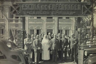 Fotografia histórica em preto e branco, tirada em frente à entrada da antiga Escola de Enfermagem "Carlos Chagas". No topo da imagem, há uma placa grande com o nome da instituição, ladeada por dois símbolos em forma de cruz estilizada. Embaixo da placa, um grupo de aproximadamente quinze pessoas posa para a fotografia, a maioria composta por homens de terno e gravata, muitos deles segurando chapéus nas mãos. Ao centro, destaca-se uma mulher vestida com uniforme branco de enfermeira, incluindo touca e luvas, sorrindo levemente. O grupo está em frente a um prédio de arquitetura clássica, com janelas e sacadas decoradas. Há automóveis antigos parcialmente visíveis nas laterais da imagem.