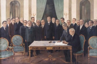 Pintura histórica de Gentil Garcez que retrata a solenidade de sanção do projeto de criação da Universidade de Minas Gerais (UMG). No centro da composição, o presidente do estado de Minas Gerais, Antônio Carlos de Andrada, está sentado à direita, assinando um documento sobre uma mesa nobre, de madeira trabalhada com detalhes dourados. Ao seu lado esquerdo, um secretário ou assessor escreve ou organiza papéis. Atrás deles, em pé, há um grupo de aproximadamente vinte homens, todos vestidos formalmente com ternos escuros e gravatas. As expressões são sérias e respeitosas, evidenciando a formalidade do momento. O ambiente é luxuoso: o salão tem paredes claras, grandes espelhos arqueados, poltronas estofadas em verde com acabamento dourado e pesadas cortinas verde-escuras ao fundo. Dois candelabros dourados com velas estão presos às paredes.