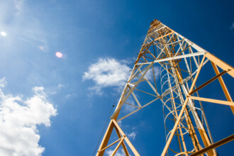A imagem mostra uma torre de transmissão alta, de estrutura metálica em treliça, vista de baixo para cima, o que enfatiza sua altura e inclinação em direção ao céu. A torre é predominantemente amarela ou laranja em suas hastes principais, com algumas seções de metal que parecem ser brancas ou prateadas, formando uma geometria complexa. No interior da estrutura da torre, é visível uma escada em espiral que sobe por toda a extensão. O fundo da imagem é um céu azul intenso, com algumas nuvens brancas esparsas. O sol é visível no canto superior esquerdo, criando um brilho intenso e algumas lentes de flare.