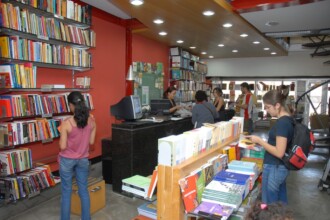 A imagem retrata o interior de uma livraria com prateleiras repletas de livros coloridos. Na parte esquerda da foto, uma mulher de blusa rosa e calça jeans está de costas para a câmera, examinando os livros em uma estante vertical. No centro da imagem, um balcão escuro com um computador e uma monitora de frente para a câmera. Há pessoas em pé no balcão e ao redor dele. À direita, outra mulher de óculos e mochila preta está de perfil, olhando para os livros dispostos em um expositor horizontal. O teto possui iluminação embutida.