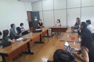 A imagem mostra um grupo de nove pessoas reunidas em uma sala de reunião com mesas de madeira dispostas em formato de "U" e cadeiras em volta. As pessoas estão sentadas, algumas usando máscaras faciais de proteção. Sobre as mesas, há garrafas de álcool em gel, copos e laptops abertos. As paredes da sala são brancas com detalhes em preto, e uma porta escura pode ser vista ao fundo. O chão é de madeira clara e o teto possui iluminação fluorescente.