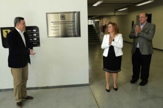 A imagem mostra um evento de inauguração ou apresentação em um ambiente interno. No centro, uma placa de metal brilhante está afixada em uma parede clara. A placa contém o brasão da "UNIVERSIDADE FEDERAL DE MINAS GERAIS" no topo e, abaixo, o texto "CENTRO DE ATIVIDADES DIDÁTICAS (CAD2)", seguido por informações sobre gestões e seus reitores. À esquerda da placa, um homem de camisa azul, blazer escuro e calça clara está em pé, gesticulando em direção à placa. À direita da placa, uma mulher de blazer branco e vestido preto está aplaudindo, e ao seu lado, um homem de óculos e blazer claro também está aplaudindo. No lado esquerdo da imagem, uma sinalização de parede com setas e números de salas é visível. O piso é de cerâmica clara, e ao fundo, uma escada pode ser vista.