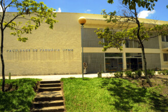 A imagem mostra a fachada de um edifício institucional de cor bege claro, que abriga a "FACULDADE DE FARMÁCIA UFMG", conforme indicado por uma inscrição em relevo na parede esquerda. A arquitetura do prédio inclui grandes janelas de vidro com divisões em grade, e uma entrada principal visível ao centro. No primeiro plano, há um caminho com degraus de pedra ou cimento que atravessa um gramado verde, subindo em direção à entrada do edifício. Duas árvores com folhagem verde emolduram a cena, uma de cada lado. Um poste de iluminação alto com uma luminária esférica amarela está posicionado na frente do prédio. O céu é azul claro com algumas nuvens brancas.