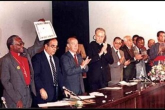 A imagem mostra uma mesa de autoridades em um evento formal. Nela, há pelo menos 10 pessoas em pé atrás de uma longa mesa de madeira escura. A maioria é branco e está batendo palmas. As pessoas usam trajes formais, como ternos e vestes religiosas, e uma delas (um homem de pele negra com colar clerical e camisa vermelha) segura um diploma com inscrições que não estão totalmente legíveis. Há microfones e papéis sobre a mesa, e à direita, um arranjo de flores. O fundo da imagem é neutro, com uma parede clara e uma divisória escura ao centro.
