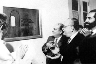 A imagem em preto e branco captura um evento de inauguração ou celebração. No centro da cena, há uma placa retangular fixada em uma parede clara. A placa, de cor escura e com borda clara, exibe a inscrição "CENTRO CULTURAL UFMG" em letras maiúsculas. Abaixo do título, há um texto menor, também em letras maiúsculas, com detalhes sobre o centro cultural. Ao lado esquerdo do texto, um logotipo abstrato com a representação de um arco está presente. À direita da placa, quatro homens são visíveis. Três deles estão no centro da imagem, e um quarto homem, com barba, aparece no lado direito, apenas com a parte superior de seu corpo visível. O homem mais próximo da placa está aplaudindo, assim como o homem mais ao fundo no lado esquerdo da imagem, cuja silhueta pode ser vista no canto inferior esquerdo.