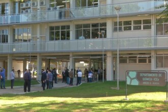 A imagem mostra a fachada de um edifício institucional moderno, com uma entrada central proeminente. O prédio é predominantemente de cor bege, com elementos em tons de bordô nas janelas e na grande estrutura triangular da entrada, que se projeta para fora. As janelas são retangulares e emolduradas com caixilhos escuros, algumas com brises ou venezianas. Um caminho pavimentado em zigue-zague leva à porta de entrada principal, que é de vidro. Há áreas de gramado verde e palmeiras decorativas nos arredores do edifício. Um poste de iluminação alto está localizado no lado direito da imagem. O céu é claro e esbranquiçado, com poucas nuvens visíveis.
