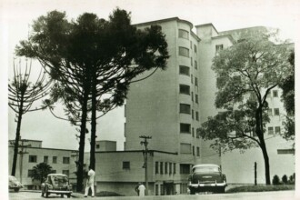 Fotografia em preto e branco tirada na década de 1960, mostrando a fachada do Hospital São Vicente de Paulo, atual Hospital das Clínicas da UFMG. O edifício possui vários andares, linhas retas e janelas verticais distribuídas de forma simétrica. Sua arquitetura é característica do estilo modernista institucional do período. Na frente do prédio, há uma via asfaltada com alguns veículos da época estacionados, incluindo carros de design arredondado. Pessoas circulam pela calçada e em frente ao prédio — algumas trajando roupas brancas, possivelmente funcionários da área da saúde. A cena é emoldurada por árvores altas. Há também postes de energia e iluminação pública que cruzam o espaço urbano.