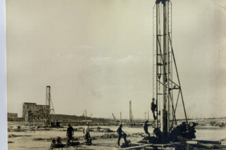 A imagem em preto e branco mostra uma vasta área de construção em um terreno irregular, sob um céu claro com algumas nuvens dispersas. Em primeiro plano, à direita, destaca-se uma estrutura alta e vertical, aparentemente de metal, com um homem subindo por uma escada acoplada a ela. Outros dois homens estão próximos à base dessa estrutura, com equipamentos ao redor. No centro da imagem, mais três homens estão visíveis no terreno. Ao longo do horizonte e em diferentes pontos da área, é possível ver várias outras estruturas verticais similares, algumas parecendo guindastes ou torres de perfuração. No fundo, à esquerda, um edifício de vários andares está em construção ou em fase de acabamento, com andaimes visíveis em sua estrutura. O terreno é irregular e parece estar sendo trabalhado.