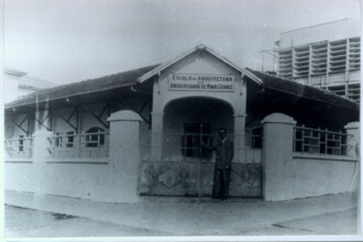 A imagem em preto e branco mostra a fachada de um edifício que abriga a "Escola de Arquitetura da Universidade de Minas Gerais", conforme indicado por uma placa sobre a entrada central. O prédio possui um telhado inclinado de telhas e paredes de cor clara. A entrada principal é um arco, com um portão de metal fechado à frente. À direita da entrada, um homem vestido com terno e sapato social está parado, de frente para a câmera. O edifício tem várias janelas laterais, algumas com grades. No lado direito e ao fundo da imagem, há um prédio mais alto com diversas janelas e o que parece ser andaimes. A área em frente ao prédio é um piso claro, e o céu é visível ao fundo.