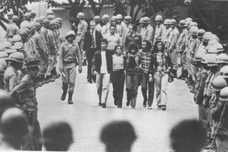 A imagem em preto e branco mostra um grupo de cinco jovens, três homens e duas mulheres, caminhando pelo centro. Eles estão vestidos com roupas casuais, como camisas xadrez, calças jeans e uma camisa social. Um dos homens está de braços dados com uma das mulheres. Eles são ladeados por duas fileiras de homens fardados e com capacetes, que parecem ser militares ou policiais, formando um corredor. No fundo, à esquerda, há um homem de terno e gravata caminhando. O ambiente geral é de um local aberto, possivelmente uma rua ou pátio.