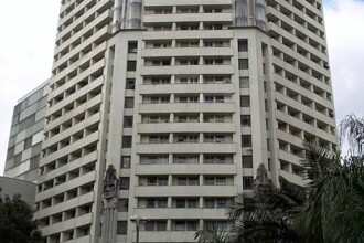 A imagem vertical colorida retrata o Edifício Acaiaca, um arranha-céu imponente localizado em Belo Horizonte, Minas Gerais. O prédio domina a composição, estendendo-se do meio inferior ao topo da imagem. Sua fachada é predominantemente clara, com múltiplas fileiras de janelas retangulares que se alinham horizontalmente em cada andar, criando um padrão repetitivo.