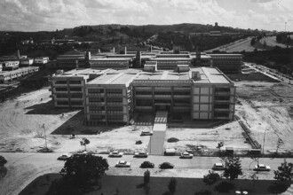 A imagem em preto e branco exibe uma vista aérea da ufmg com vários edifícios modernistas e amplas áreas de terreno, alguns arborizados e outros com terra aparente. No centro, um grande complexo de edifícios horizontais, com múltiplas janelas e telhados planos, é interligado por passarelas. Em primeiro plano, carros de modelo antigo estão estacionados em uma área pavimentada. Árvores jovens e vegetação rasteira são visíveis em várias partes do campus. Ao fundo, uma paisagem montanhosa se estende com mais edificações distantes, e o céu está parcialmente nublado.