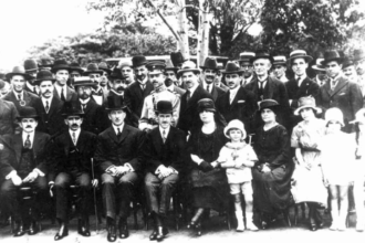 A imagem em preto e branco mostra um grande grupo de pessoas, predominantemente homens, posando para uma foto em um ambiente externo com árvores ao fundo. A maioria dos indivíduos veste trajes formais, como ternos escuros e chapéus. Na primeira fila, quatro homens estão sentados em cadeiras, e entre eles e à direita, há algumas mulheres e crianças, também em poses formais. A luz do sol incide sobre o grupo, criando algumas sombras. A vegetação ao fundo é densa, com folhagem escura.