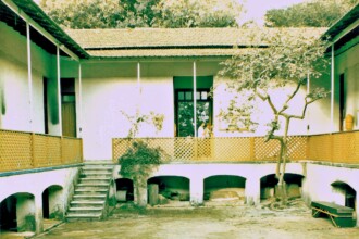 A imagem colorida mostra o pátio interno de um casarão antigo, visto de um ângulo levemente elevado. O pátio é quadrado ou retangular, cercado por paredes claras e uma varanda no segundo andar. A varanda possui um guarda-corpo de madeira com desenhos vazados em tom mostarda e é sustentada por pilares brancos. O telhado do casarão, visível na parte superior central, é de telhas claras. No centro da imagem, há uma escadaria de alvenaria com degraus de pedra que leva do pátio para a varanda do segundo andar. Abaixo da varanda, há uma série de arcos que parecem ser aberturas para um subsolo ou áreas de depósito, com algumas sombras e objetos indistintos em seu interior. No lado direito do pátio, um portão fechado de madeira em tom escuro está visível. Uma árvore de tronco fino e galhos espalhados, com pouca folhagem, está no canto direito do pátio. No lado esquerdo, a varanda continua, e no fundo, é possível ver a vegetação de árvores altas.