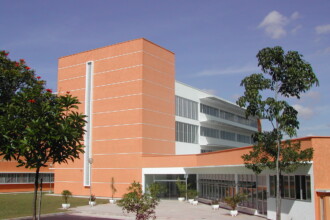 A imagem exibe a fachada de um edifício moderno e multifacetado, com predominância de paredes em tom laranja pastel e seções brancas. A estrutura apresenta um bloco vertical proeminente na cor laranja com uma faixa vertical branca. A entrada principal, visível na parte inferior central, é caracterizada por grandes painéis de vidro. O edifício possui várias janelas horizontais e brises que se estendem ao longo de algumas seções. Árvores com folhagem verde e um gramado bem cuidado adornam o entorno da construção. O céu, em um tom de azul claro, está parcialmente visível acima do edifício.