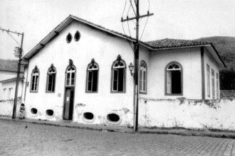 Fotografia em preto e branco de um prédio histórico de arquitetura colonial, localizado em uma rua de paralelepípedos. Trata-se da antiga sede da Faculdade Livre de Direito. A construção tem fachada assimétrica e janelas altas com arcos ogivais, decoradas com molduras ornamentais. O muro branco que circunda parte do prédio apresenta sinais de desgaste. À esquerda da imagem, vê-se uma pessoa próxima ao muro. Um poste de madeira e fios elétricos cortam parte da vista frontal do edifício. Ao fundo, há uma paisagem com morros.