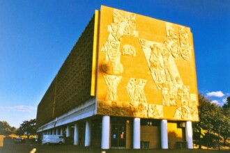 A imagem colorida retrata o prédio da Reitoria da UFMG em Belo Horizonte, um edifício de arquitetura modernista com uma fachada marcante. A lateral direita do prédio, voltada para o observador, apresenta um grande painel artístico em relevo, com figuras humanas e elementos simbólicos em tons de amarelo e ocre. A fachada principal, à esquerda, possui uma série de pequenas janelas retangulares dispostas em um padrão quadriculado, com brises verticais. O edifício é elevado do solo por uma série de pilares cilíndricos brancos, criando um espaço aberto no nível térreo. Em frente ao prédio, há uma área gramada com algumas árvores e veículos estacionados ao fundo, incluindo uma van branca. O céu acima está azul claro com algumas nuvens brancas esparsas, e a iluminação sugere um dia ensolarado.
