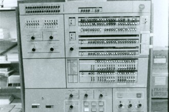 A imagem em preto e branco exibe de perto um computador IBM System/360, uma máquina de grande porte característica das décadas de 1960 e 1970. O painel de controle do computador domina o centro da imagem, preenchido com múltiplos mostradores, botões, luzes indicadoras e interruptores, organizados em seções retangulares. Na parte superior do painel, os logotipos "IBM" e "System 360" são visíveis, com a palavra "PENSE" centralizada abaixo. À esquerda e à direita do computador, estantes com pilhas de livros ou documentos são visíveis, e caixas com a palavra "FRÁGIL" impressa podem ser vistas no lado direito. A iluminação é uniforme, sugerindo um ambiente interno.