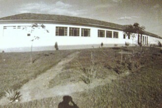 A imagem em sépia mostra um edifício longo e horizontal com telhado de telhas escuras, visto de um ângulo baixo que enfatiza o terreno em primeiro plano. O prédio possui uma fachada clara com múltiplas janelas retangulares, algumas em grupos, que se estendem por sua extensão. A construção parece ter um único andar visível na frente. Em primeiro plano, um gramado com grama curta e algumas trilhas de terra que se curvam. Pequenos arbustos e árvores jovens estão espalhados pelo gramado, e uma planta de folhagem espessa é visível no canto inferior esquerdo. Uma sombra escura, possivelmente de quem tirou a foto, é visível no centro inferior da imagem. O céu na parte superior da imagem está claro, com algumas nuvens dispersas.