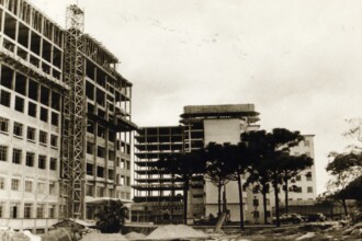 A imagem em preto e branco retrata um cenário de construção urbana, com edifícios de diferentes estágios de conclusão e algumas árvores em primeiro plano. À esquerda, um grande edifício está em construção avançada, exibindo sua estrutura de concreto aparente e múltiplas janelas em seus muitos andares. Andaimes metálicos estão visíveis na lateral, acompanhando a altura da construção. Há pilhas de materiais de construção na base do prédio. Ao centro e à direita, outro edifício, possivelmente parte do mesmo complexo, é visível. Uma parte está em fase de estrutura, com andares e colunas de concreto expostos, enquanto a outra parece mais concluída, com fachadas e janelas aparentes. Algumas árvores, incluindo pinheiros, estão em frente a esse prédio. Carros de modelo antigo estão estacionados em uma rua ou área pavimentada no lado direito da imagem. O céu está claro e a iluminação é uniforme.