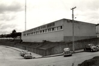 A imagem em preto e branco mostra a fachada de um edifício que abriga a "ESCOLA DE EDUCAÇÃO FÍSICA DE MINAS GERAIS", conforme inscrito na parte superior da parede frontal. O prédio, de arquitetura robusta e linhas retas, possui uma longa fileira de janelas horizontais que se estendem por quase toda a sua largura. Em primeiro plano, uma rua pavimentada e uma área de estacionamento à esquerda contam com três carros antigos estacionados: dois carros de modelo sedã mais próximos da câmera e dois Fuscas à direita, na parte inferior da imagem. Um poste de madeira com fiação e uma luminária está visível à direita do edifício, enquanto uma torre de rádio ou antena alta se eleva no fundo, à esquerda do prédio. O céu está parcialmente nublado.
