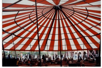 A imagem mostra o interior de uma grande tenda circular com um telhado de lona listrado em vermelho e branco, alternando as cores em segmentos que convergem para o centro superior. Estruturas metálicas escuras formam a armação da tenda, com pilares verticais e vigas que se curvam para sustentar a lona. No chão da tenda, há um grande número de pessoas reunidas, algumas sentadas em degraus ou arquibancadas, e outras em pé. A parte inferior da tenda é aberta, permitindo vislumbrar o exterior com árvores e algumas construções ao fundo.