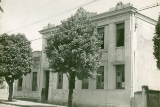 A imagem em tons sépia retrata a fachada de um edifício de dois andares. A construção apresenta uma arquitetura simétrica, com janelas retangulares e altas no segundo andar e uma série de janelas e uma porta no térreo. A parte superior do edifício possui um acabamento ornamentado, com detalhes em relevo acima das janelas do andar superior e no topo da platibanda. Em frente ao prédio, há uma calçada e uma rua. Duas árvores com folhagem densa estão posicionadas em primeiro plano, uma à esquerda e outra, maior e centralizada, em frente à fachada do edifício. Fios de energia atravessam a parte superior da imagem. À direita, um muro baixo e um pilar de concreto marcam o limite da propriedade. A iluminação da cena sugere um dia claro.