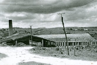A imagem em preto e branco mostra uma construção industrial em um cenário rural. No lado esquerdo da imagem, há uma chaminé alta e estreita de tijolos, indicando a presença de uma fábrica. Anexado à chaminé, um telhado inclinado e escuro cobre parte da estrutura, que parece ser um galpão. Em frente a esta seção, uma pilha de material escuro está no chão, possivelmente carvão ou outro insumo. À direita do galpão inicial, estende-se um longo edifício horizontal com um telhado de telhas claras. Este edifício possui várias janelas retangulares alinhadas. Postes de madeira com fios atravessam o primeiro plano e o meio da imagem, sugerindo linhas de transmissão elétrica ou telefonia. Ao fundo, o terreno é ondulado com vegetação esparsa e algumas estruturas de casas distantes são visíveis sob um céu com nuvens. O chão no primeiro plano é de terra batida, com algumas áreas de vegetação rasteira.