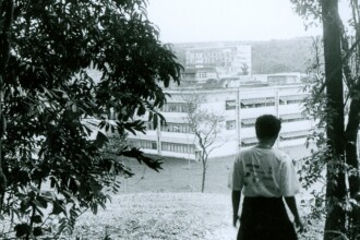 A imagem em preto e branco apresenta uma pessoa de costas, vista de um ponto de vista elevado, observando um complexo de edifícios ao longe. A pessoa, cujo gênero não pode ser determinado, veste uma camiseta de cor clara e uma calça escura, e está no centro inferior da imagem. O primeiro plano é emoldurado por folhagem escura de árvores nas laterais. Um caminho em declive leva a um terreno gramado. Ao fundo, destacam-se dois edifícios de múltiplos andares, com janelas horizontais e brises. O edifício mais próximo é menor e fica em um plano inferior ao edifício mais distante, que parece ser maior e mais complexo em sua estrutura. O céu é claro e uniforme.