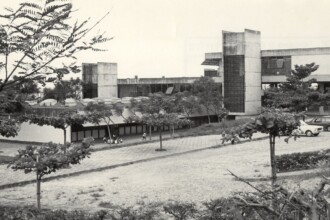 A imagem em preto e branco apresenta uma vista ampla de um complexo institucional, caracterizado por edifícios com arquitetura modernista e grandes áreas verdes. O edifício principal, visível no centro e à direita, possui múltiplos andares com janelas horizontais e elementos de concreto aparente, incluindo grandes pilares verticais e platibandas. Em primeiro plano, há um extenso gramado e árvores de pequeno a médio porte com folhagem dispersa. No lado direito da imagem, uma área de estacionamento exibe vários carros de modelos antigos. A iluminação é difusa, sugerindo um dia nublado ou com luz suave.