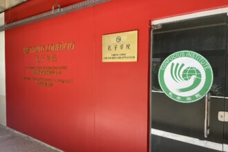 A imagem mostra a entrada de um instituto em um edifício com paredes vermelhas vibrantes. À esquerda, uma placa dourada na parede exibe em relevo "INSTITUTO CONFÚCIO" em português e, abaixo, caracteres chineses. Mais abaixo, há outro texto em português e chinês, de menor tamanho. No centro da parede vermelha, outra placa dourada, menor, traz o texto "孔子学院 Confucius Institute at the Federal University of Minas Gerais". À direita, uma porta de vidro tem um adesivo circular grande e branco com borda verde. Dentro do círculo, há um logotipo em verde que representa uma mão segurando um globo, e também o nome "CONFUCIOUS INSTITUTE" em semicírculo na parte superior e caracteres chineses na parte inferior. O chão é cinza claro, e a iluminação parece ser artificial, vinda de cima.