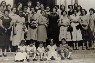 A imagem em preto e branco retrata um grupo de 26 mulheres e 5 crianças, posando para uma fotografia em uma escadaria externa de um edifício. As mulheres estão dispostas em várias fileiras sobre os degraus, com as crianças sentadas nos degraus mais baixos em primeiro plano. As mulheres vestem trajes formais da época, como vestidos longos, saias, blusas e casacos, com penteados variados. As crianças também estão bem-vestidas, com roupas claras. O fundo da imagem mostra a entrada de um edifício, com uma porta escura e detalhes arquitetônicos simples. A iluminação da cena é uniforme, sem sombras pronunciadas, sugerindo um dia claro.