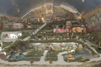 A imagem colorida apresenta um grande e elaborado presépio, ocupando toda a extensão do quadro. A cena representa a cidade de Belém e a Natividade em uma paisagem montanhosa e detalhada, com numerosas figuras humanas e animais em miniatura, além de construções e elementos naturais simulados. O presépio é iluminado por várias fontes de luz, incluindo um candelabro no topo e luzes coloridas que destacam diferentes áreas da cena, como casas, rios e montanhas. O céu simulado acima é escuro com estrelas e uma representação da aurora. A composição inclui rios sinuosos, pontes, caminhos, vegetação artificial e uma gruta que provavelmente abriga a cena do nascimento de Jesus, embora essa área específica não esteja totalmente visível.