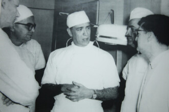 A imagem em preto e branco mostra um grupo de cinco homens em um ambiente que parece ser um centro cirúrgico ou sala de exames. Todos os homens usam uniformes médicos brancos e toucas cirúrgicas brancas. O homem no centro da imagem está de frente para o observador e tem as mãos unidas à frente do corpo, enquanto fala. Um relógio de pulso escuro é visível em seu braço esquerdo. Os outros quatro homens estão ao redor dele, em diferentes posições, aparentemente atentos ao que ele diz. Ao fundo, alguns equipamentos médicos, como uma luminária de teto, podem ser parcialmente vistos.